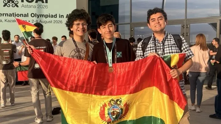 Santiago Soto, Nicolás Bravo con su medalla y el entrenador Diego Angulo sostienen la bandera de Bolivia frente a la sede de la IOAI en Astana