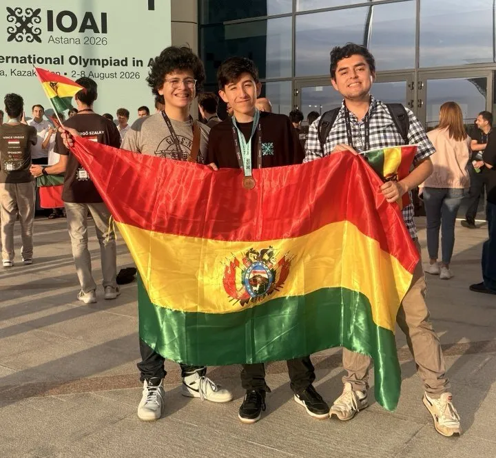 Santiago Soto, Nicolás Bravo con su medalla y el entrenador Diego Angulo sostienen la bandera de Bolivia frente a la sede de la IOAI en Astana