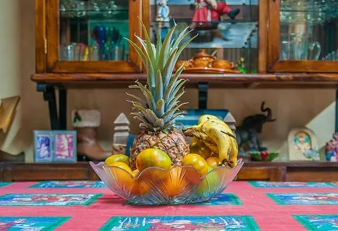 Un frutero de vidrio con una piña, naranjas y plátanos sobre una mesa con mantel de colores.