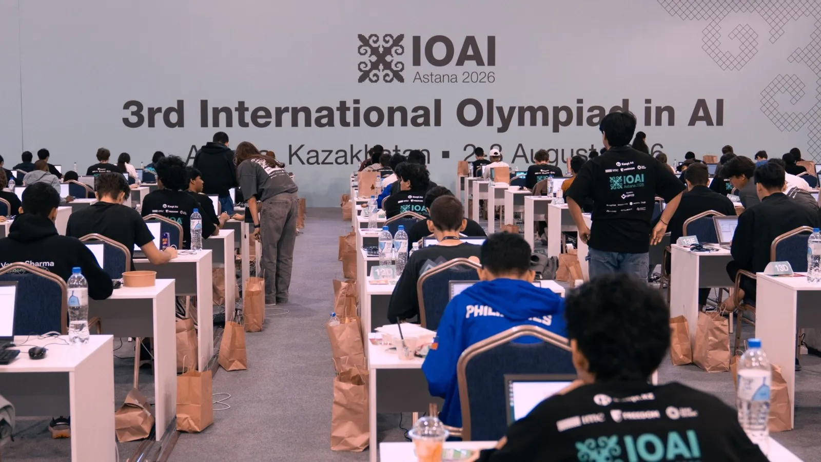 Sala de la prueba: decenas de estudiantes frente a sus computadoras bajo el cartel de la tercera Olimpiada Internacional de IA